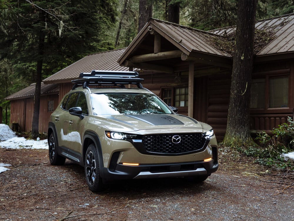 ¾ shot of Zircon Sand Metallic Meridian trim CX-50 parked 7/8 front on in front of log cabin in the forest.  