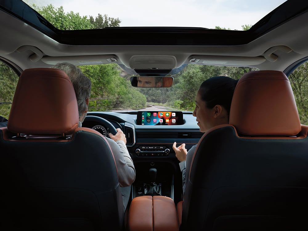 From 2nd row seats looking forward, a man drives a CX-50 down a wintery road, a smiling woman in the passenger seat is looking at him. 