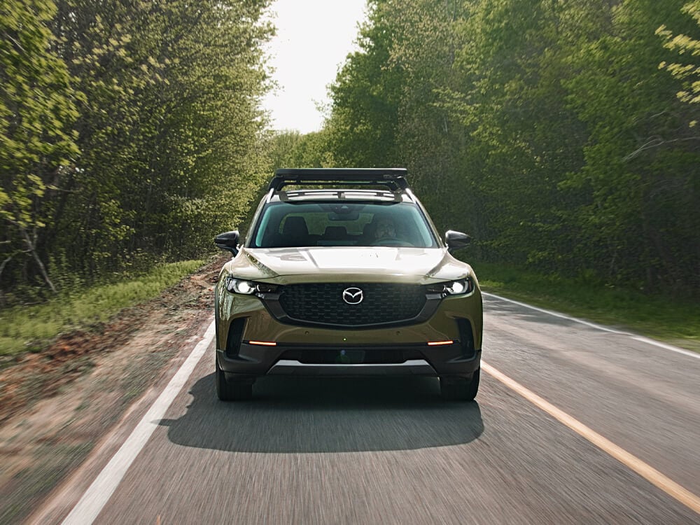 A Zircon Sand Metallic CX-50 is driving straight towards camera on paved road in the forest. 