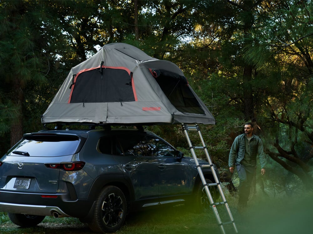 Ingot Blue Metallic CX-50 with roof rack with pitched tent and ladder on roof, parked on dirt and grass in forest. An outdoorsy man is walking away from the vehicle. 