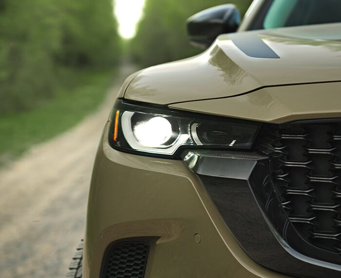 Close up of headlight of Zircon Sand Metallic CX-50 driving toward camera on dirt/gravel road.