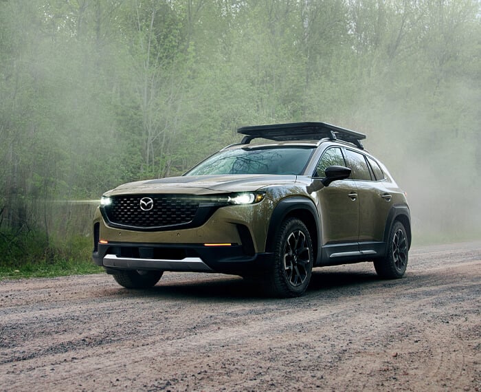 Un CX-50 sable de zircon métallisé vu de ¾ avec les phares allumés, circulant en forêt. 