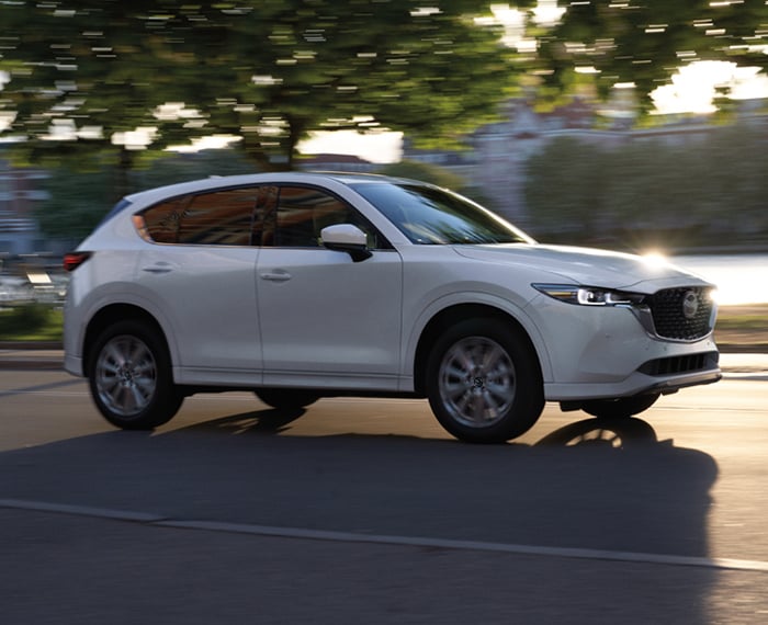 Plan de profil d’un CX-5 blanc rhodium qui circule avec des bâtiments majestueux et des arbres flous en arrière-plan. 