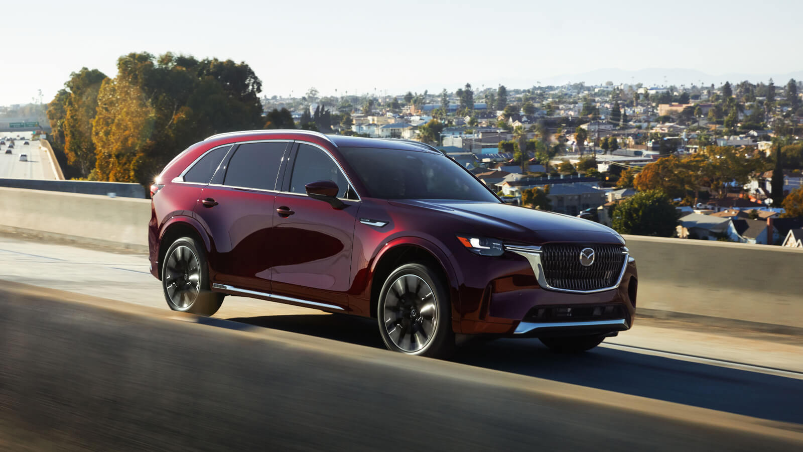 Artisan Red CX-90 drives on a viaduct, sun reflecting on its side. A suburban area is in the background.