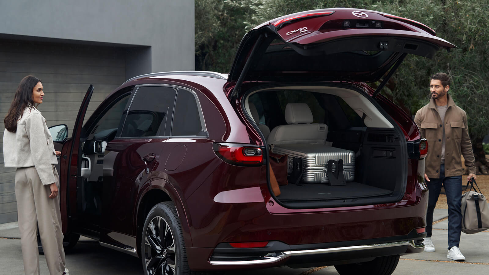 Couple loading Artisan Red CX-90 with back liftgate open revealing a large silver suitcase. Man walks towards rear with a bag, woman opens driver’s side door.