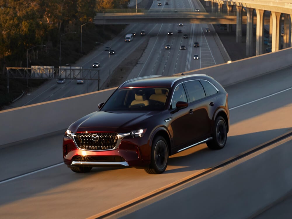 Artisan Red CX-90 PHEV drives toward sunset on concrete highway overpass.