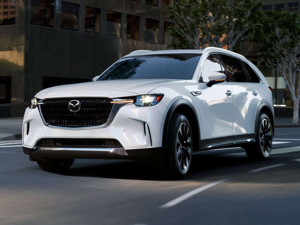 Vue en contreplongée de ¾ face d’un CX-90 blanc arctique dans une courbe le long d’un boulevard urbain bordé d’immeubles majestueux avec des arbres en arrière-plan. 