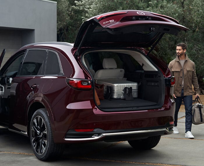 Couple loading Artisan Red CX-90 with back liftgate open revealing a large silver suitcase. Man walks towards rear with a bag, woman opens driver’s side door.