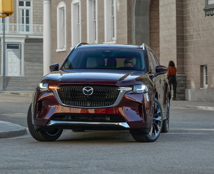 Artisan Red CX-90 almost straight grille on, making a turn in a city block.