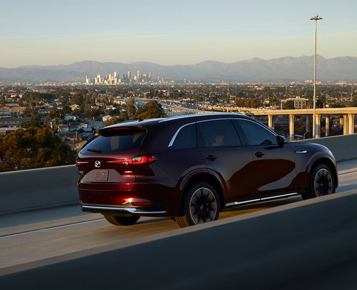 Artisan Red CX-90 drives away from sunset on concrete highway overpass.