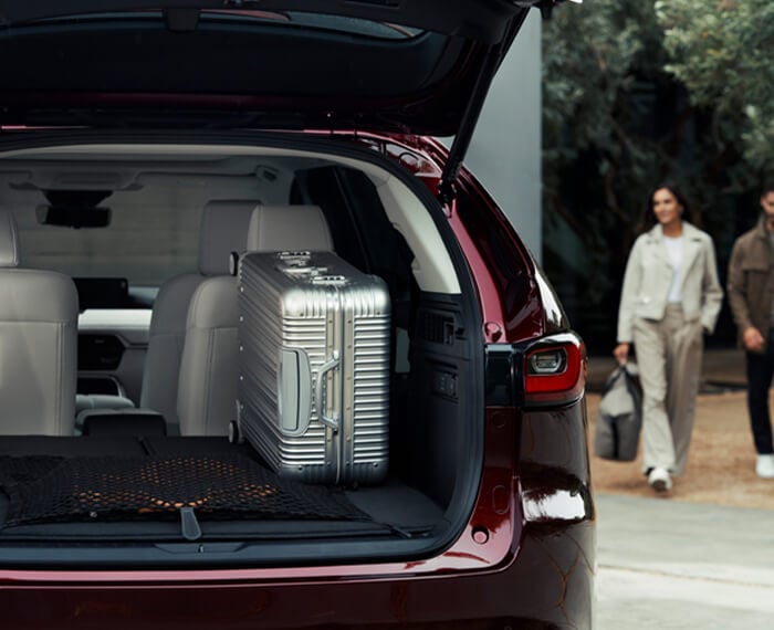 Couple approaches Artisan Red CX-90 with back lift gate open revealing a large silver suitcase. 