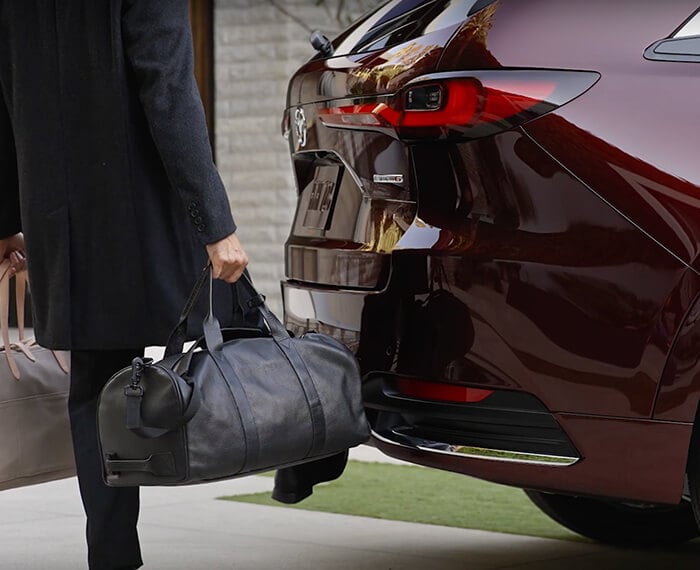 Un homme aux mains pleines utilise son pied pour activer le hayon électrique mains libres d’un CX-90 rouge artisan. 