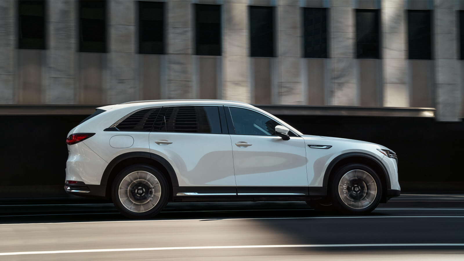 Vue de profil d’un CX-90 hybride rechargeable blanc arctique passant devant un édifice en béton minimaliste et moderne. 