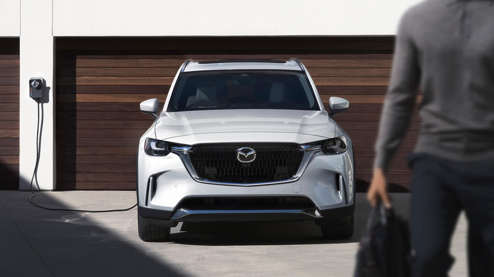 Un VÉHR CX-90 blanc arctique, branché et en mode recharge, garé devant un garage double avec des portes de garage à lattes de bois.