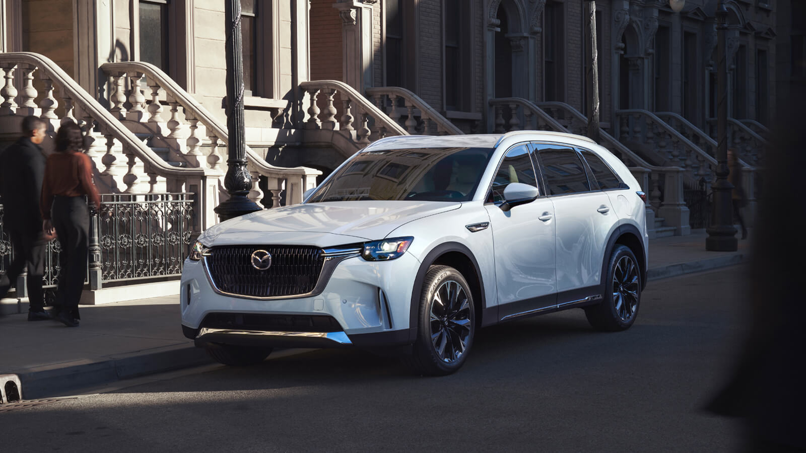 Un CX-90 blanc arctique garé dans une rue urbaine bordée d’escaliers d’appartements en béton. 