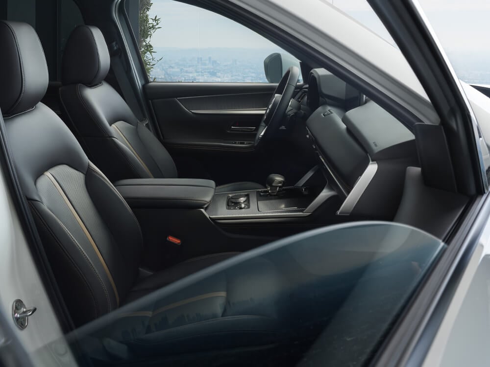 Par la fenêtre partiellement ouverte, côté passager, vue sur les deux sièges avant, le tableau de bord et la console. On distingue une ville au loin à travers la vitre du côté conducteur. 