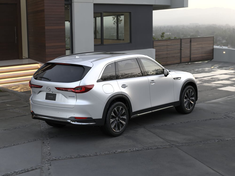 En arrière-plan, en plongée de ¾, un CX-90 blanc arctique est garé en biais devant une maison moderne et élégante.   