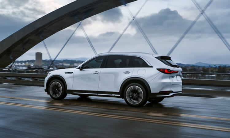 7/8 profile shot of Arctic White CX-90 Plug-In Hybrid driving on a bridge, motion blur on road in foreground. 