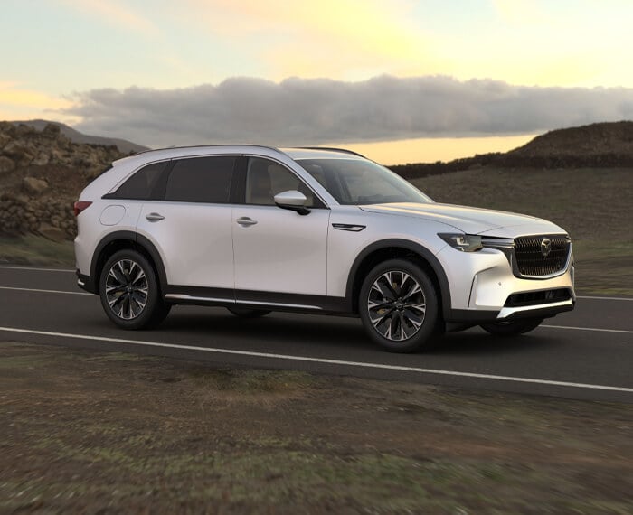 ¾ front shot of Arctic White CX-90 driving through hilly countryside at dawn/dusk.