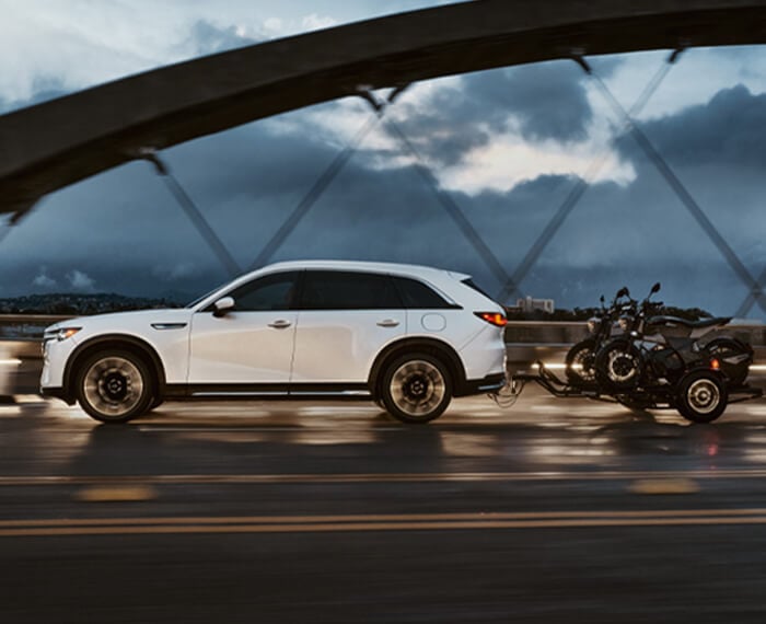 Un CX-90 hybride rechargeable blanc arctique traversant un pont au crépuscule en tirant une petite remorque avec deux motos. 