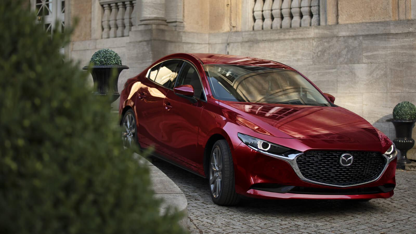 Soul Red Crystal Metallic Mazda3 sedan front ¾ shot, parked on cobblestone drive in front of a limestone historical building. 