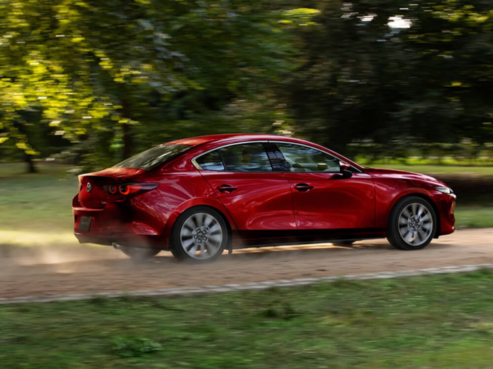 Soul Red Crystal Metallic Mazda3 sedan drives swiftly along country road kicking up dust, passing large trees in the background. 