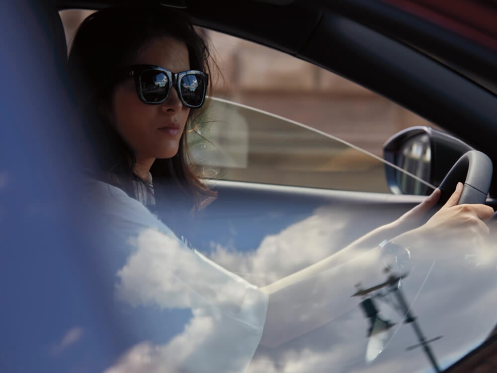 De la fenêtre du passager avant, on voit une conductrice assise au volant d’une berline Mazda3. Des nuages et le ciel se reflètent dans la vitre. 