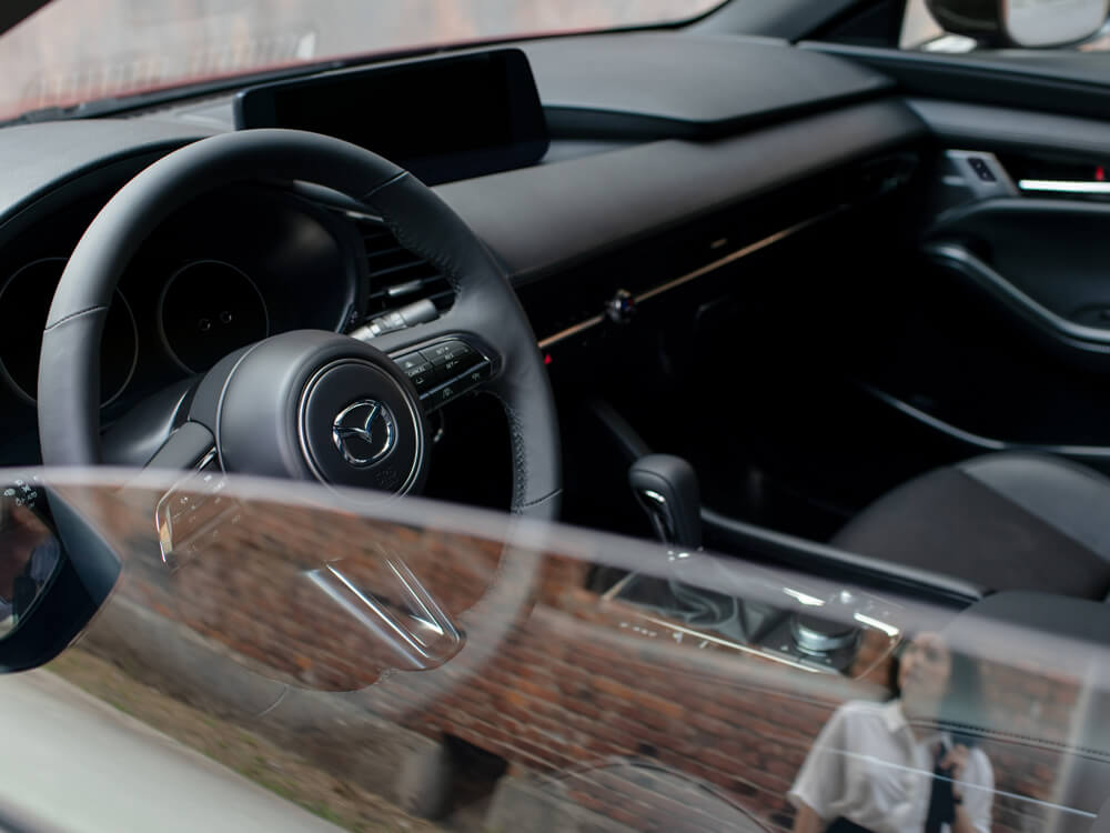 L’intérieur d’une berline Mazda3 vu de la fenêtre côté passager avant, avec le reflet du conducteur qui s’approche dans la vitre à moitié baissée. 