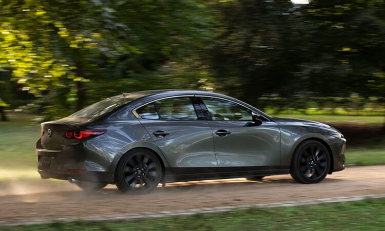 Mazda3 sedan drives swiftly along cobblestone drive kicking up dust, passing trees in the background. 