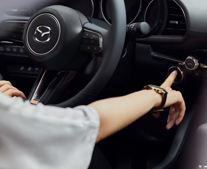 Close-up of steering wheel and front driver interior of Mazda3 Sedan, the driver’s hand is pressing the engine Start button. 
