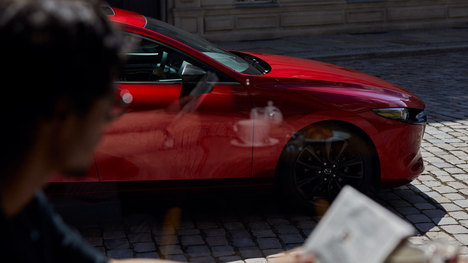 Gros plan du devant d’une Mazda3 Sport rouge vibrant cristal métallisé garée dans une rue pavée.