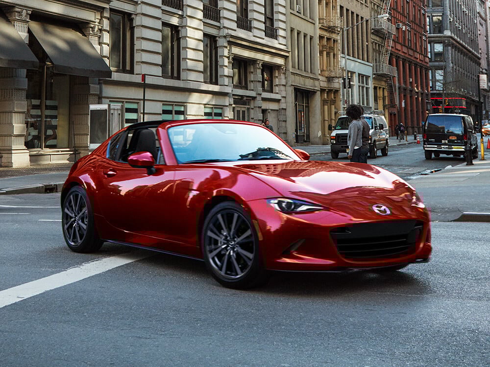 3/4ths view of a red MX-5 RF turning down a city street with buildings and pedestrians in the background.