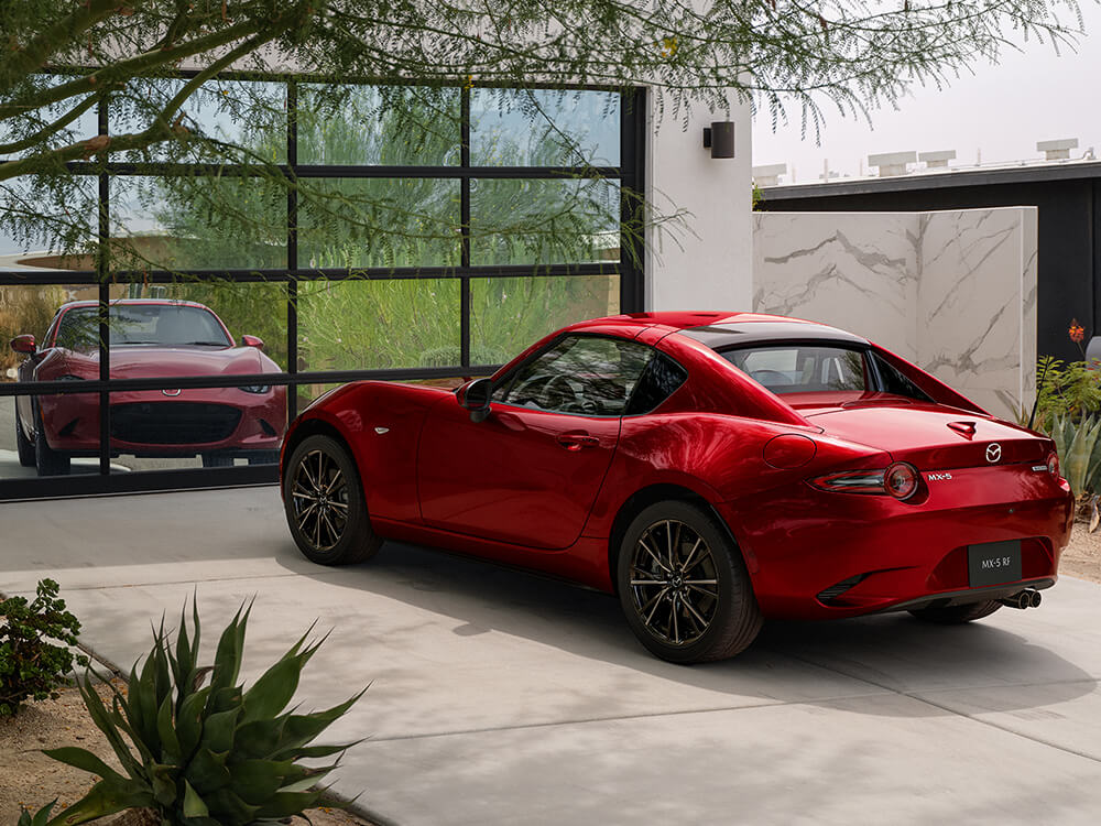 A red MX-5 RF with its top up sits parked in a driveway, reflected in the glass of a large, multi-paneled window.