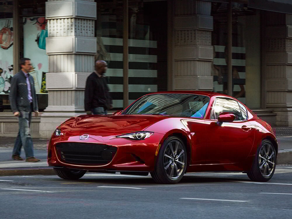 A red MX-5 RF with its top up drives down a city street with buildings and two pedestrians visible in the background.