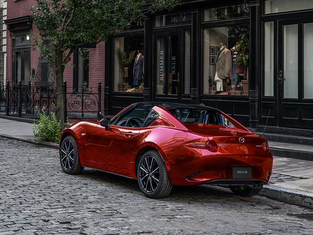 View of the back of a red MX-5 RF with its top down parked on a city street in front of a clothing store.