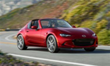 A red MX-5 RF drives down a winding road surrounded by hilly terrain. 