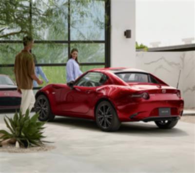 Deux personnes regardent une MX-5 RF rouge garée devant une vitrine miroitante.