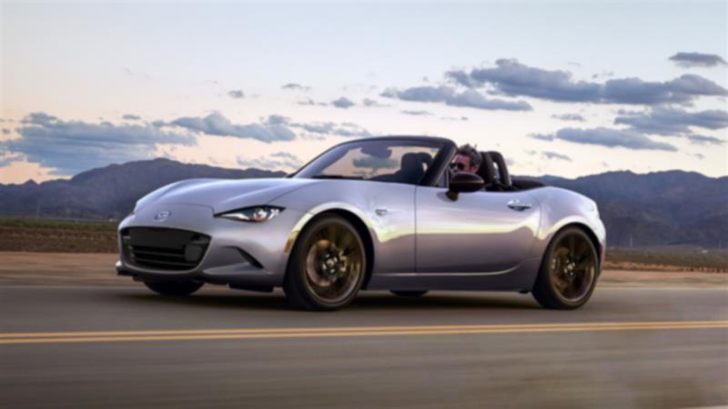 Une MX-5 ST avec son toit ouvert roule sur une route dégagée près de montagnes.