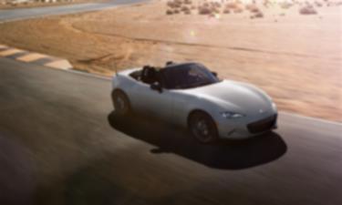 Une MX-5 ST avec son toit ouvert roule sur une route désertique près d’un terrain sablonneux.