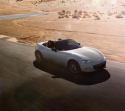 Une MX-5 ST avec son toit ouvert roule sur une route désertique près d’un terrain sablonneux.