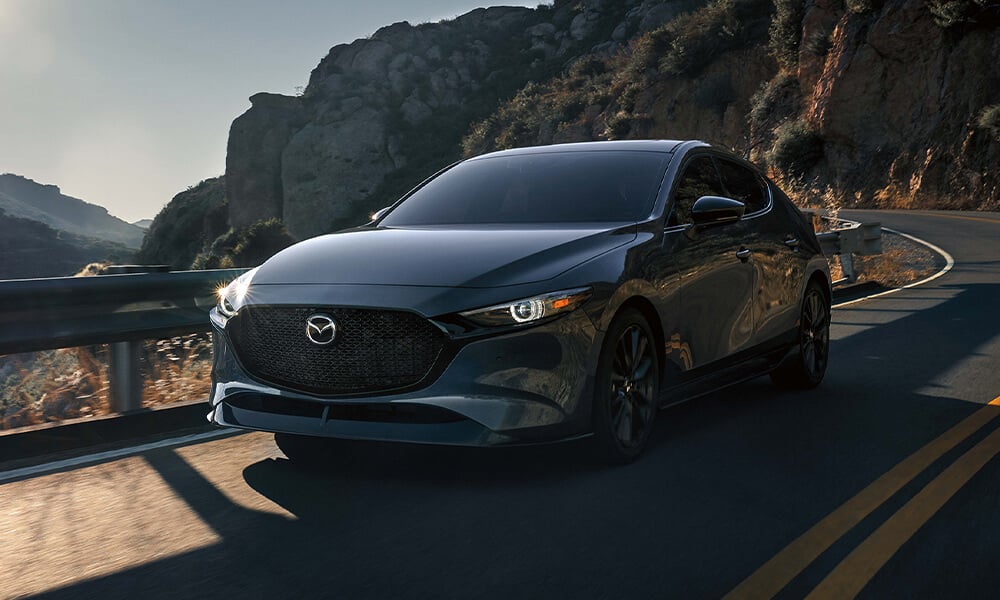 Front view of a Deep Crystal Blue Mica Mazda3 Sport driving on a winding highway.
