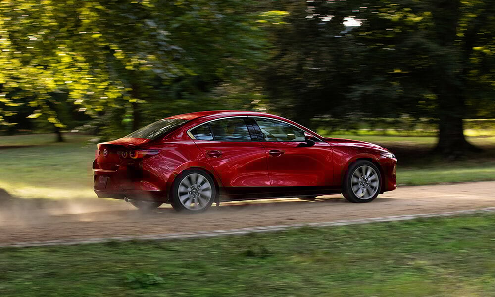 Side profile of a Soul Red Crystal Metallic Mazda3 driving on a dirt road, kicking up dust.