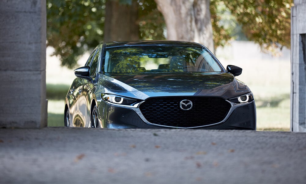 Front view of a Deep Crystal Blue Mica Mazda3, with the front wheels partially obscured by the road, appearing to drive up a hill.